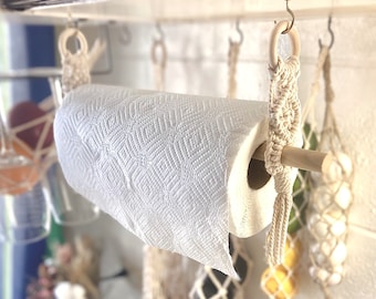 Puede incluir: Un rollo de papel de cocina blanco con un patrón de diamantes está sujeto por un soporte de papel de cocina de macramé. El soporte está hecho de cuerda de color natural y una clavija de madera. El soporte cuelga de un gancho de metal.