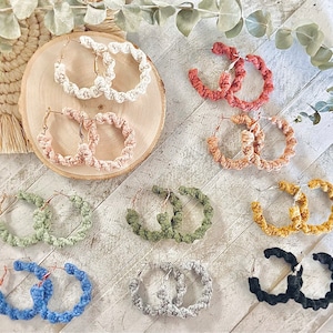 May include: Assortment of 10 pairs of handmade crocheted hoop earrings in various colors, including beige, brown, green, gray, and black. The earrings are displayed on a light wood surface.