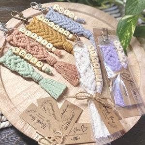May include: A set of six personalized keychains with wooden beads and macrame tassels. The keychains are in various colors, including pink, yellow, green, white, and purple. Each keychain has a different name spelled out on the beads. The keychains are packaged in clear plastic bags with brown paper tags that say "Thank You" and "Will you help me tie the knot?"