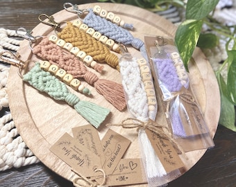 May include: A collection of macrame keychains in light blue, mustard yellow, dusty rose, sage green, white, and purple. Each keychain features wooden beads spelling names like COCO, SOPHIA, OLIVIA, LOGAN, HARPER, and EMMA, finished with a metal clasp.