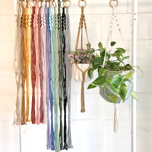 May include: A collection of macrame plant hangers in various colors including beige, yellow, orange, pink, purple, blue, green, black, and grey. Each hanger features a wooden ring at the top and a knotted cord design with a tassel at the bottom.