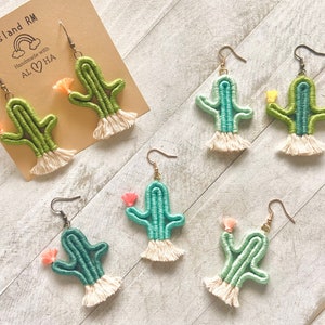 Pode incluir: Um conjunto de seis brincos de cacto feitos à mão. Os brincos são feitos de fio verde e azul e têm uma franja de fio branco. Os brincos são decorados com pequenas flores rosa e amarelas.