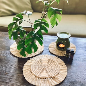 Op de afbeelding: Drie macramé-onderzetters met franjes, één met een rond midden en twee met een vierkant midden. Een plant in een pot met groene bladeren staat op een van de onderzetters. Een groene keramische olieverdamper met een kaars erin staat op een andere onderzetter. Twee kleine flesjes etherische olie staan op de tafel.