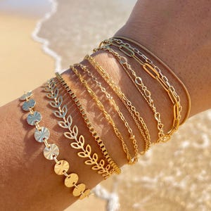 May include: A stack of gold bracelets on a person's wrist. The bracelets are made of different chain styles, including a chain with small leaves, a chain with small circles, and a chain with small links.