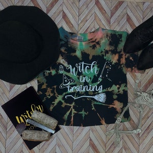 May include: A black tie-dye t-shirt with the words "Witch in training" in white, a black witch hat, black boots, a skeleton, a black journal with gold lettering, and sage bundles. The shirt has green, orange, and pink accents.