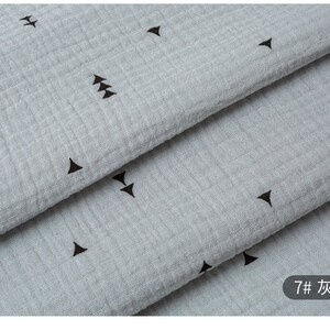 May include: Close-up of a light gray fabric with a subtle texture and a repeating pattern of small black triangles. The fabric is folded in layers, revealing the pattern.