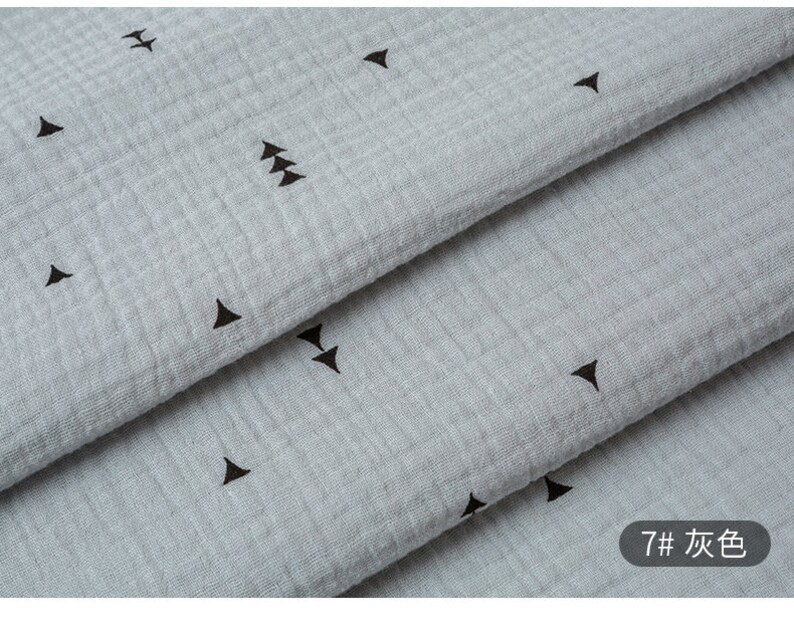 May include: Close-up of a light gray fabric with a subtle texture and a repeating pattern of small black triangles. The fabric is folded in layers, revealing the pattern.