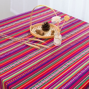 May include: A colorful striped tablecloth with a red background and multi-colored stripes. The tablecloth is decorated with small white and black geometric patterns. A wooden tray with a pine cone and a small glass bottle are on top of the tablecloth.