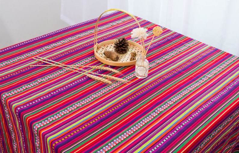 May include: A colorful striped tablecloth with a red background and multi-colored stripes. The tablecloth is decorated with small white and black geometric patterns. A wooden tray with a pine cone and a small glass bottle are on top of the tablecloth.