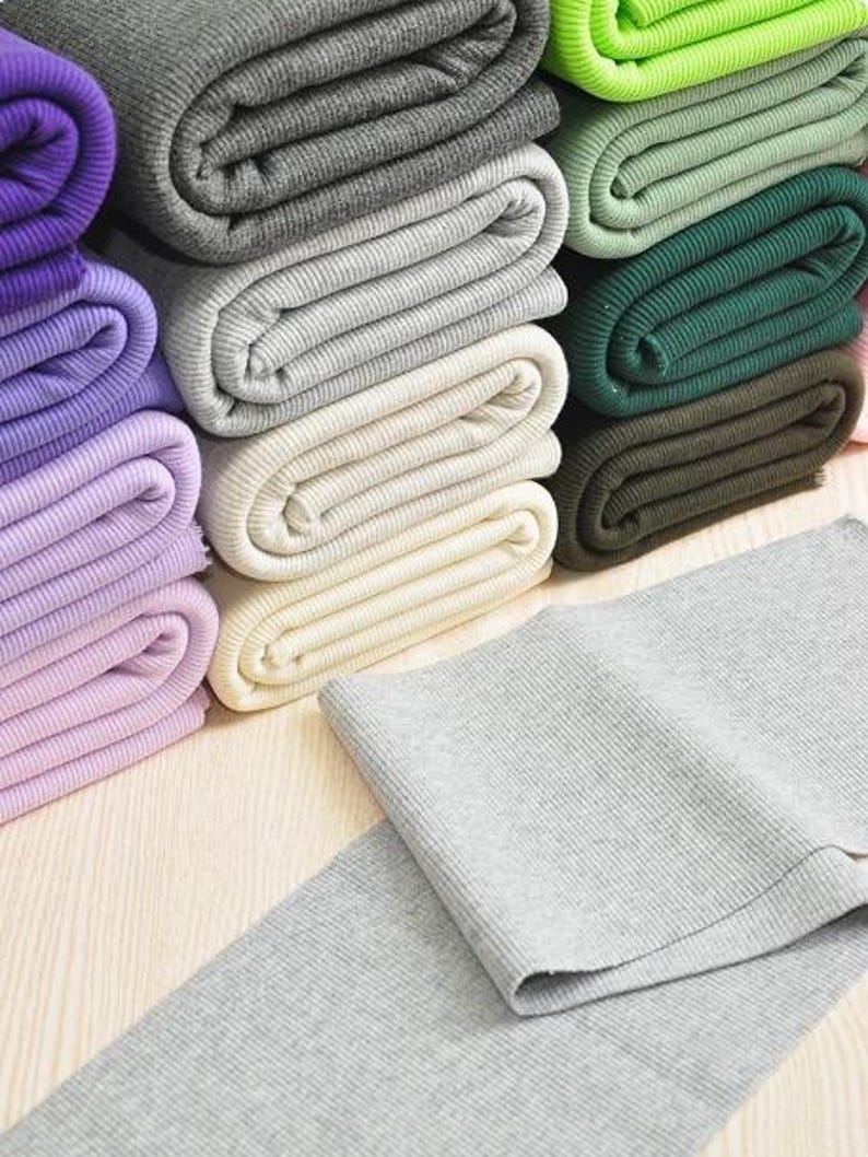 May include: A stack of different colored ribbed knit fabric rolls. The rolls are stacked in a vertical column, with the top roll being green. The rolls are all folded in half, and the fabric is visible on both sides. The fabric is a soft, stretchy material that is perfect for sewing projects.