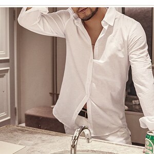 May include: A white linen shirt with long sleeves, partially unbuttoned, worn with white pants. The person is standing in front of a mirror, with a can of beer visible. The shirt has a classic collar and buttoned cuffs.