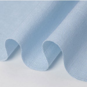 May include: Close-up of a light blue fabric with a textured weave. The fabric is draped in soft folds, creating gentle curves and shadows. The material appears to be lightweight and breathable, suitable for clothing.