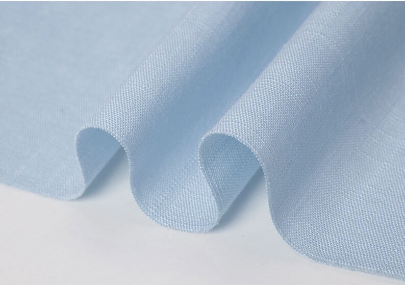 May include: Close-up of a light blue fabric with a textured weave. The fabric is draped in soft folds, creating gentle curves and shadows. The material appears to be lightweight and breathable, suitable for clothing.