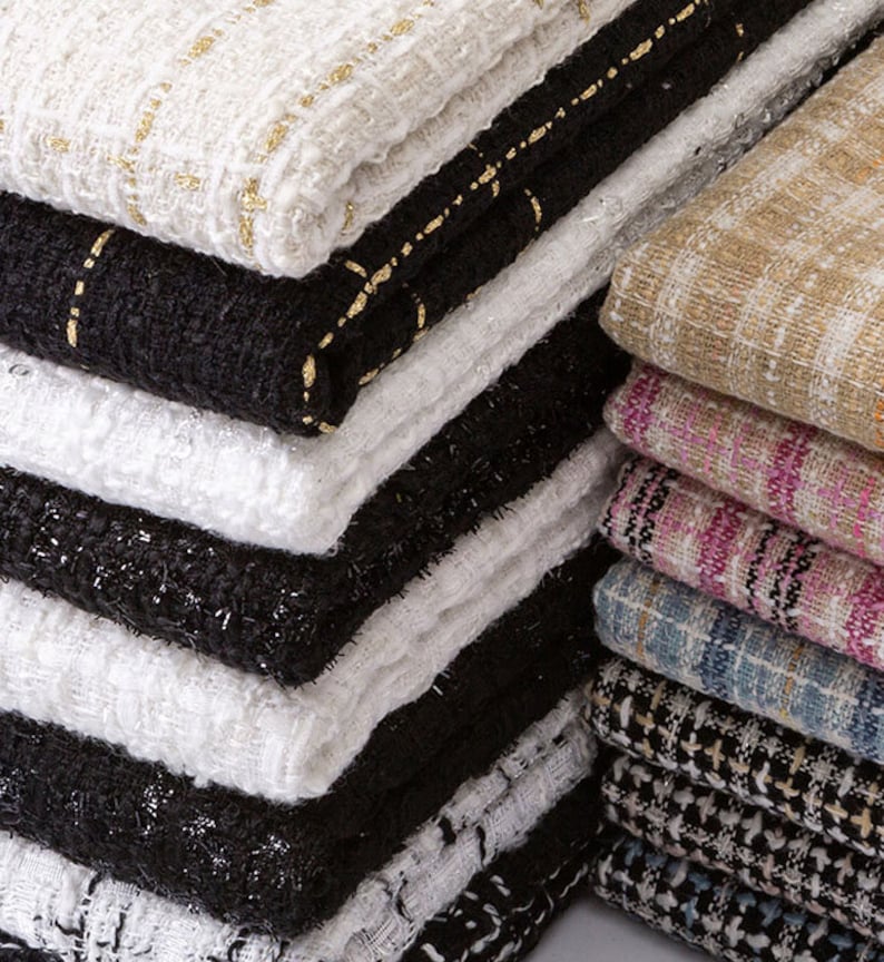 Op de afbeelding: Een stapel verschillende stoffen met patronen in verschillende kleuren, waaronder wit, zwart, beige, roze en blauw. De stoffen zijn geweven met een tweedachtige textuur en hebben een subtiele glans.