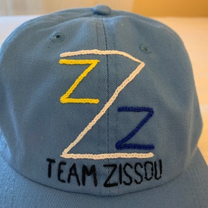 May include: A light blue baseball cap with a white, yellow, and blue embroidered 'Z' design and the text 'TEAM ZISSOU' in black embroidery.