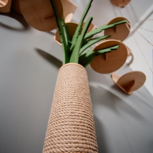 May include: A tall, brown, rope-covered scratching post with a green, leafy plant-shaped top. The post is against a gray wall.