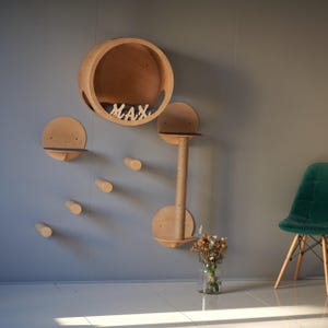 May include: A wooden cat tree with a round bed, shelves, and scratching posts. The cat tree is mounted on a gray wall and has the name "MAX" spelled out in wooden letters.