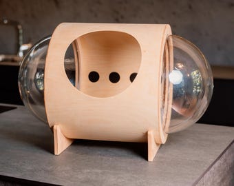 Può includere: Un letto per gatti in legno con una cupola in plastica trasparente. Il letto ha un'apertura rotonda e tre piccoli fori sul lato. Il letto è appoggiato su un piano di lavoro grigio.