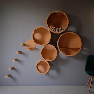 Puede incluir: Un conjunto de seis estantes circulares de madera montados en una pared gris. Los estantes están dispuestos en forma escalonada, con el estante más grande en la parte superior y el más pequeño en la parte inferior. Todos los estantes están hechos de madera natural y tienen un acabado liso. La palabra "BELLA" está escrita en letras de madera en el estante superior.