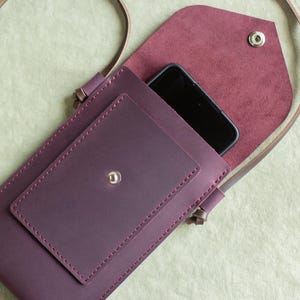 Puede incluir: Un bolso bandolera de cuero color morado oscuro para teléfono móvil con un bolsillo frontal rectangular con cierre de broche plateado, una correa larga y fina, y una solapa puntiaguda que asegura el compartimento principal con un broche metálico.