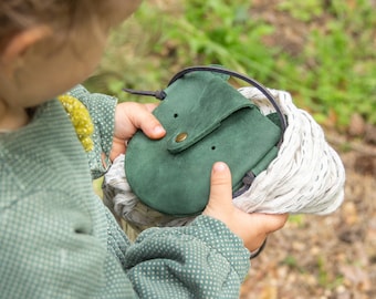 May include: A small, rounded dark green suede leather pouch featuring a flap closure secured with a single brass-colored metal snap button. The pouch has a thin dark leather strap and is nestled within a textured, off-white woven fabric wrap.