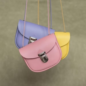 Könnte beinhalten: Drei kleine, pastellfarbene Handtaschen hängen vor einem gedämpften grünen Hintergrund. Die Handtaschen sind in den Farbtönen Lavendel, Rosa und Gelb gehalten, jede mit einem silbernen Verschluss und einem passenden Riemen. Die Taschen haben eine abgerundete Form.