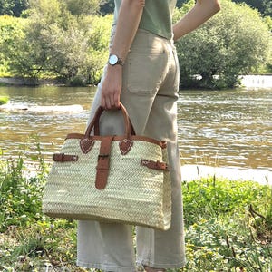 May include: A woven straw tote bag with brown leather details. The bag has a rectangular shape with a top handle and a buckle closure. The bag is held by a person standing near a river.