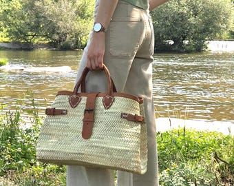 May include: A woven straw tote bag with brown leather details. The bag has a rectangular shape with a top handle and a buckle closure. The bag is held by a person standing near a river.