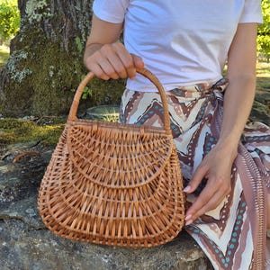 May include: A woven wicker handbag with a curved top and a handle. The bag is light brown and has a natural, rustic appearance. The bag is held by a person wearing a white t-shirt and patterned skirt, sitting on a stone wall.