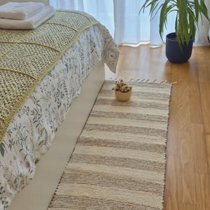 Peut inclure: Intérieur de chambre avec un lit recouvert d'une housse à motifs floraux et d'une couverture en tricot vert clair. Un tapis rayé aux tons neutres est posé sur le sol en bois. Une plante en pot et des serviettes blanches sont également visibles.