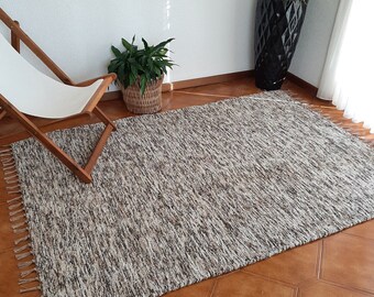 Puede incluir: Una alfombra rectangular y texturizada con una paleta de colores neutros de beige, marrón y crema. La alfombra tiene un borde con flecos y está colocada sobre un suelo de baldosas marrones. Una tumbona de madera y plantas en macetas están en el fondo.