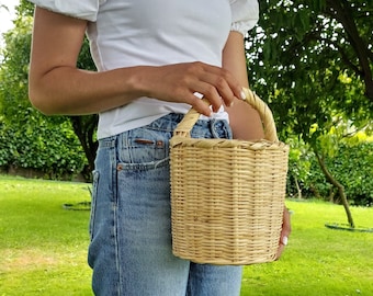 May include: A woman is holding a light brown wicker basket with a woven handle. The basket is round and has a natural finish.