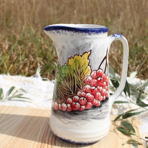 May include: A white ceramic jug with a blue rim and a handle. The jug is decorated with a hand-painted design of red berries and green leaves.