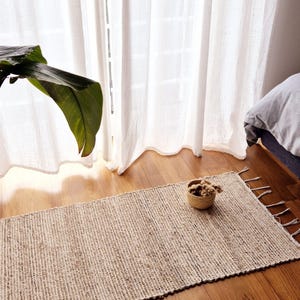 May include: A rectangular, beige woven rug with fringe detail, placed on a hardwood floor. The rug is near a window with sheer white curtains and a bed with a striped comforter. A potted plant is in the foreground.