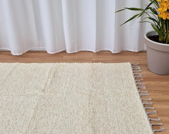 May include: Cream-colored rectangular rug with a textured weave and fringed edges. The rug is placed on a wooden floor, with a white curtain in the background. A potted plant with yellow flowers is visible.
