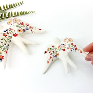 May include: Two white ceramic swallows with colorful floral designs. The swallows have intricate patterns of red, yellow, blue, and green flowers and leaves. One swallow is being held by a hand. The swallows are on a white background.