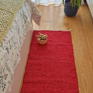 Può includere: Un tappeto rettangolare rosso vivo con una trama strutturata è posato su un pavimento in legno. Il tappeto è posizionato vicino a un letto con un copriletto a fantasia floreale e una coperta gialla intrecciata. Un piccolo cestino di fiori secchi è sul tappeto.
