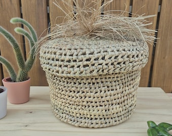 May include: A round woven basket with a lid, crafted from natural fibres. The basket has a textured surface. A small green ceramic bird figurine is placed nearby. Cacti in small pots are in the background.