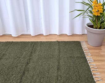 May include: An olive green rectangular rug with a textured surface and fringed edges. The rug is on a wooden floor, with a white curtain in the background. A potted plant with yellow flowers is also visible.