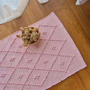 Può includere: Un tappeto rettangolare rosa con un motivo a rombi e dettagli a pois in rilievo. Un piccolo cestino intrecciato pieno di fiori secchi è appoggiato sul tappeto. Il tappeto è su un pavimento in legno.