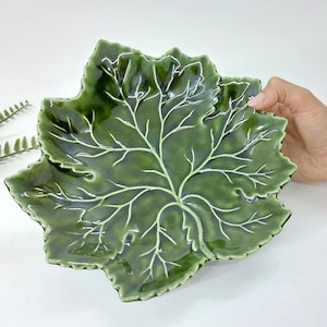 May include: A green ceramic leaf-shaped serving dish with white vein detailing. The dish has a glossy finish and is held by a hand. The leaf design is detailed and the dish is likely used for serving food or as a decorative piece.
