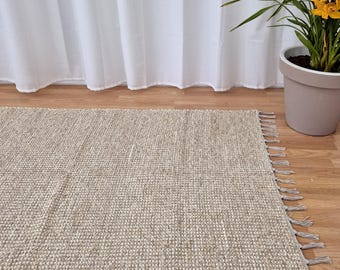 May include: A rectangular, beige woven rug with fringe detail. The rug is placed on a wooden floor, with a white curtain in the background. A potted plant with yellow flowers is to the right of the rug.