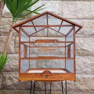Puede incluir: Una jaula de pájaros de madera con un acabado marrón y una malla de alambre. La jaula tiene un techo en forma de pico y se apoya en tres patas de horquilla negras.