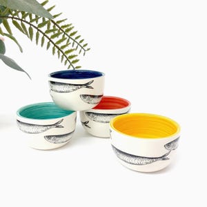 Puede incluir: Un conjunto de cuatro cuencos pequeños de cerámica con exterior blanco y dibujos de peces en líneas negras. Cada cuenco tiene un color interior distinto: azul intenso, turquesa, naranja y amarillo brillante, dispuestos en grupo.
