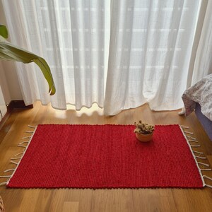 Op de afbeelding: Een rechthoekig rood tapijt met witte franjes ligt op een houten vloer. Een kleine, ronde, geweven mand met gedroogde bloemen staat op het tapijt. Transparante witte gordijnen zijn op de achtergrond.