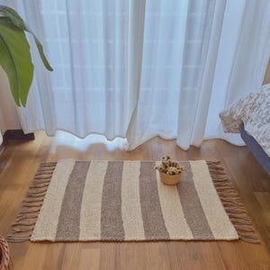 Può includere: Un tappeto rettangolare con strisce verticali beige e marroni alternate. Il tappeto ha un bordo sfrangiato ed è posto su un pavimento in legno. Un piccolo cestino intrecciato con fiori secchi è posto sul tappeto.