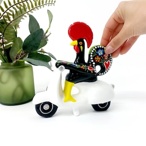 May include: A decorative ceramic rooster figurine riding a white scooter. The rooster is black with colorful floral designs, a red comb, yellow legs, and a yellow beak. The scooter has black tires and a white body.