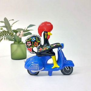 Puede incluir: Un scooter azul con una figura de gallo negro montando en él. El gallo tiene una cresta y una barbilla rojas, y sus plumas de la cola están pintadas con flores de colores.