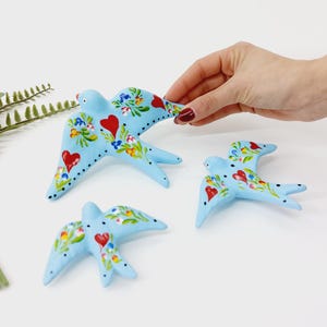 May include: Three blue ceramic birds with painted red hearts and floral designs. The birds are in flight and are a decorative home accent.