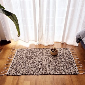 Op de afbeelding: Een rechthoekig, handgeweven vloerkleed met een gespikkeld bruin en beige patroon. Het kleed heeft franjes aan de korte uiteinden. Een klein, rond geweven mandje met gedroogde planten staat op het kleed.